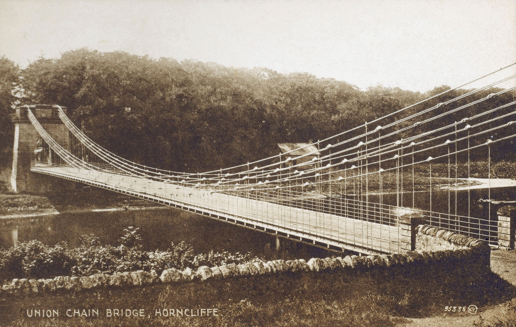 A sepia-toned picture shows the deck and piers of a bridge that spans a body of water. The suspension system comprises iron rods and chains.