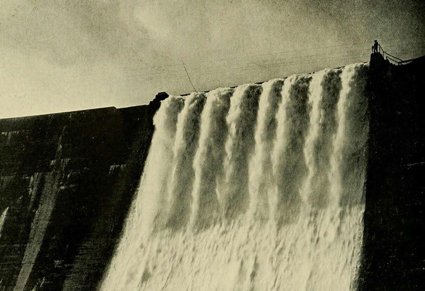 Picture shows a black and white image of water rushing over the edge of a dam.