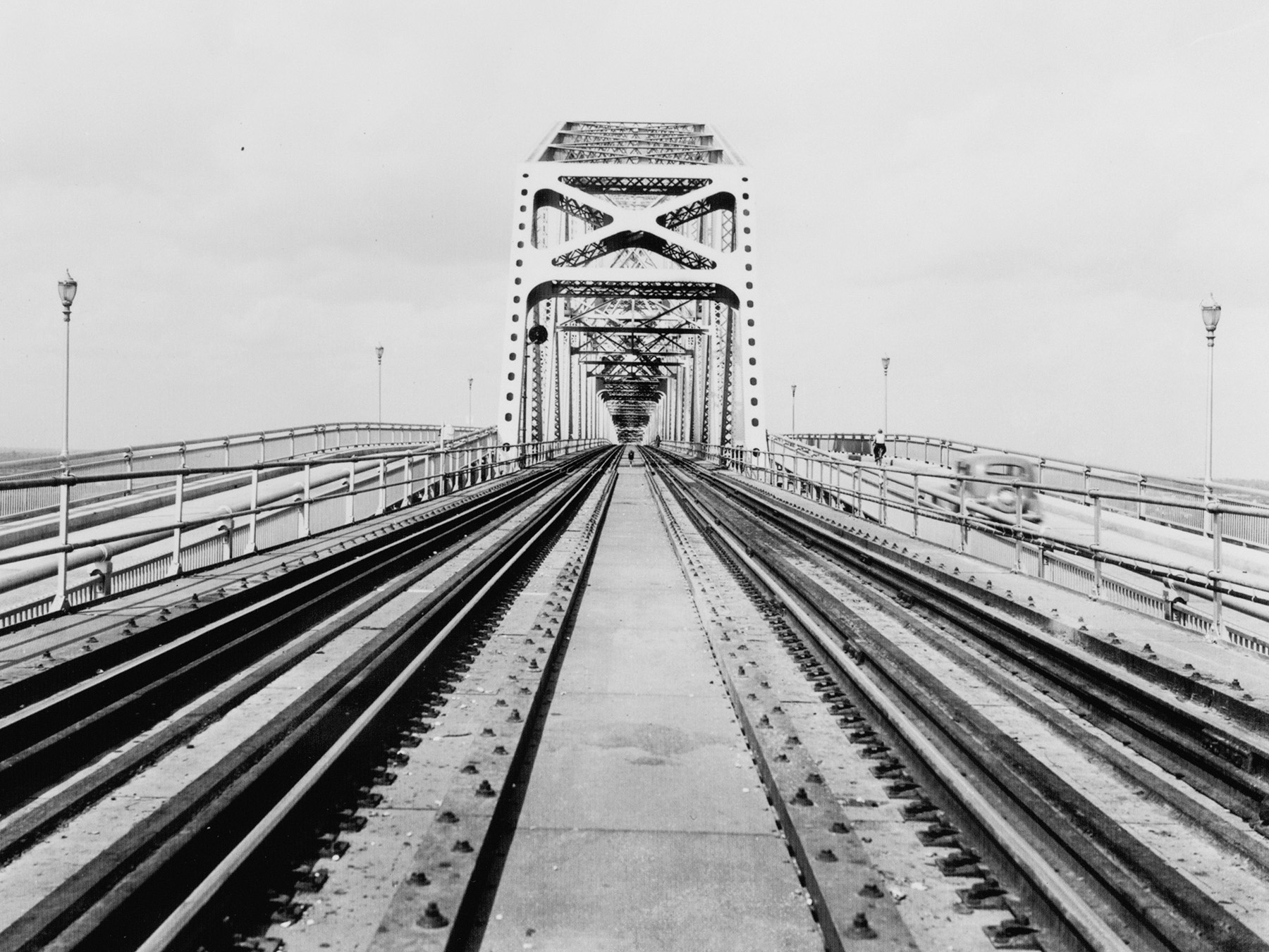 Photo shows a bridge with railroad tracks in its center and a highway on either side. A sole car is seen on the bridge.