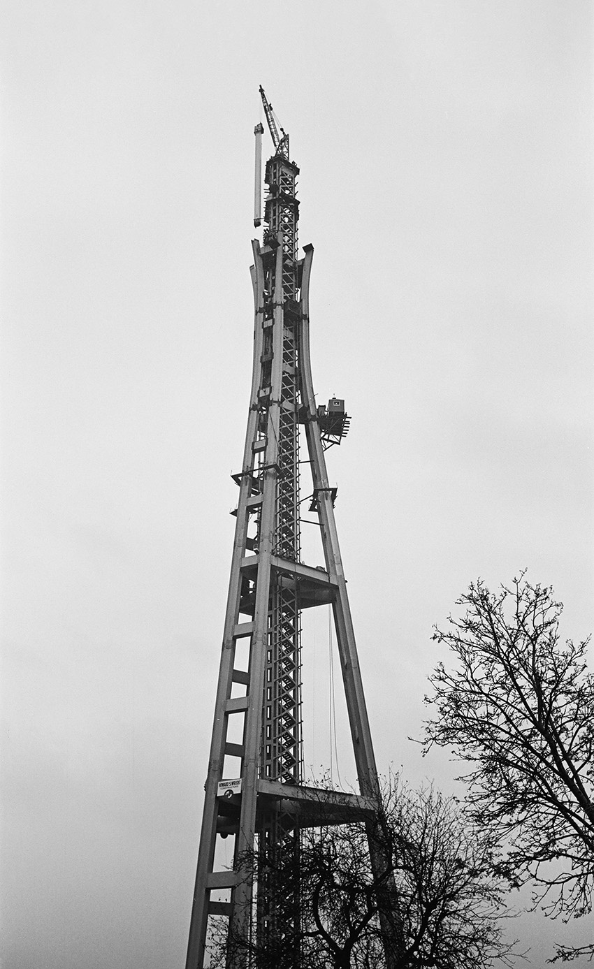 The photograph shows a tall structure with long thin, steel columns connected to a central core. A column piece is being added to the structure.