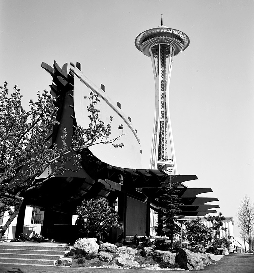 In the foreground is a one-story building. There are steps leading up to the door. IN the background is a tall, thin, steel structure known as the Space Needle. The Space Needle has three steel “legs” that are crowned by a saucer-like structure.