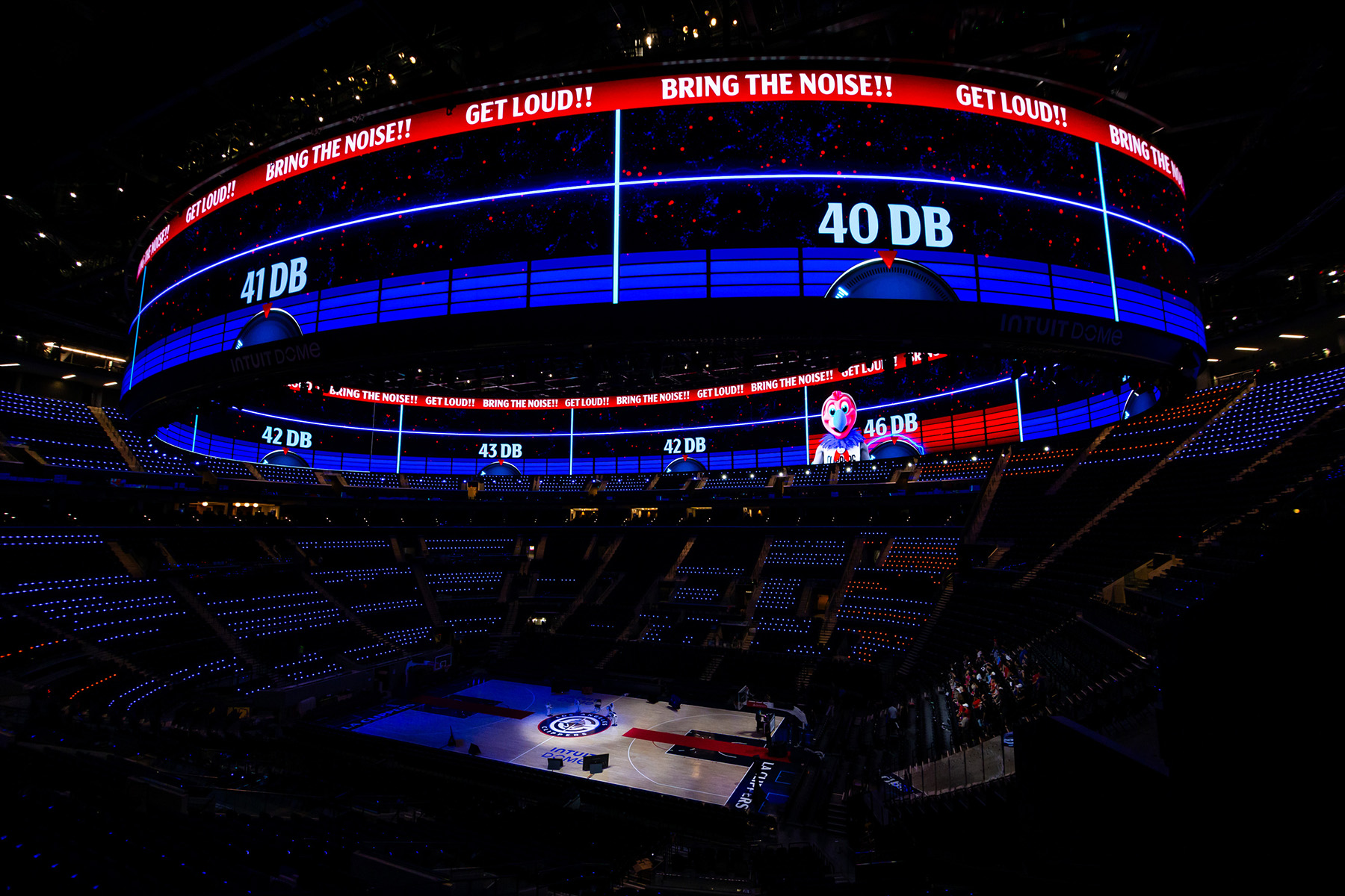 Photograph shows a giant, circular board anchored to trusses in the ceiling. The basketball court can be seen below the halo board.
