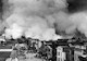 a black and white image from above shows smoke billowing in the background as people scramble in the streets among buildings